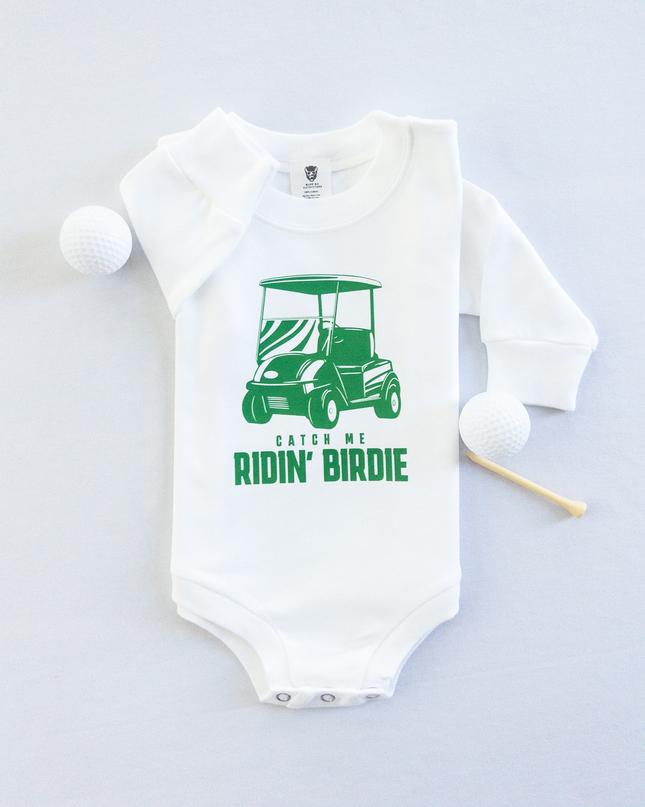 White baby golf onesie featuring a green golf cart graphic and playful text reading 'Catch Me Ridin' Birdie,' styled flat with two golf balls and a wooden tee, fun golf-themed outfit for babies and toddlers.