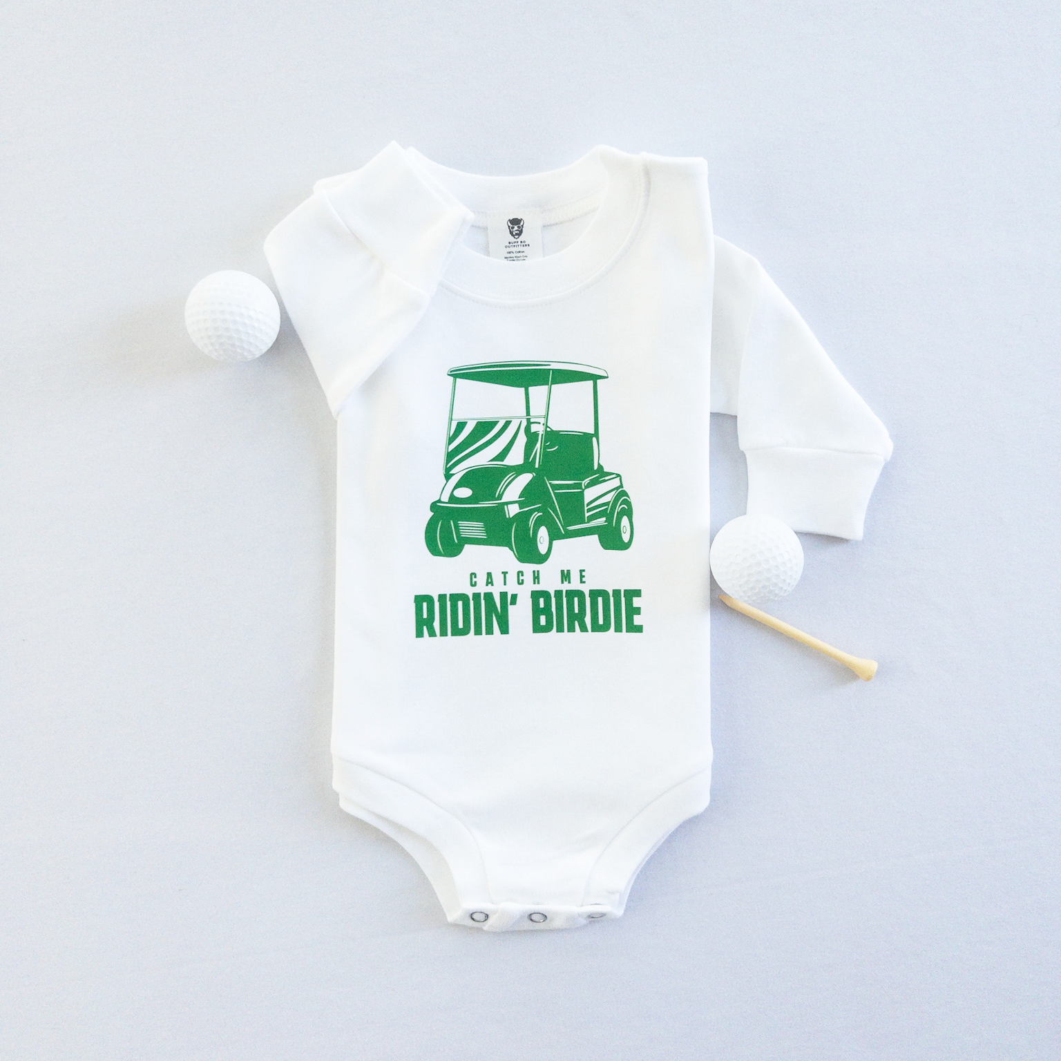 White baby golf onesie featuring a green golf cart graphic and playful text reading 'Catch Me Ridin' Birdie,' styled flat with two golf balls and a wooden tee, fun golf-themed outfit for babies and toddlers.