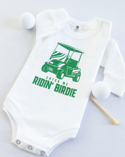 White baby golf onesie featuring a green golf cart graphic and playful text reading 'Catch Me Ridin' Birdie,' styled flat with two golf balls and a wooden tee, fun golf-themed outfit for babies and toddlers.