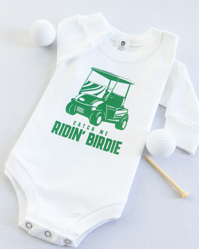 White baby golf onesie featuring a green golf cart graphic and playful text reading 'Catch Me Ridin' Birdie,' styled flat with two golf balls and a wooden tee, fun golf-themed outfit for babies and toddlers.