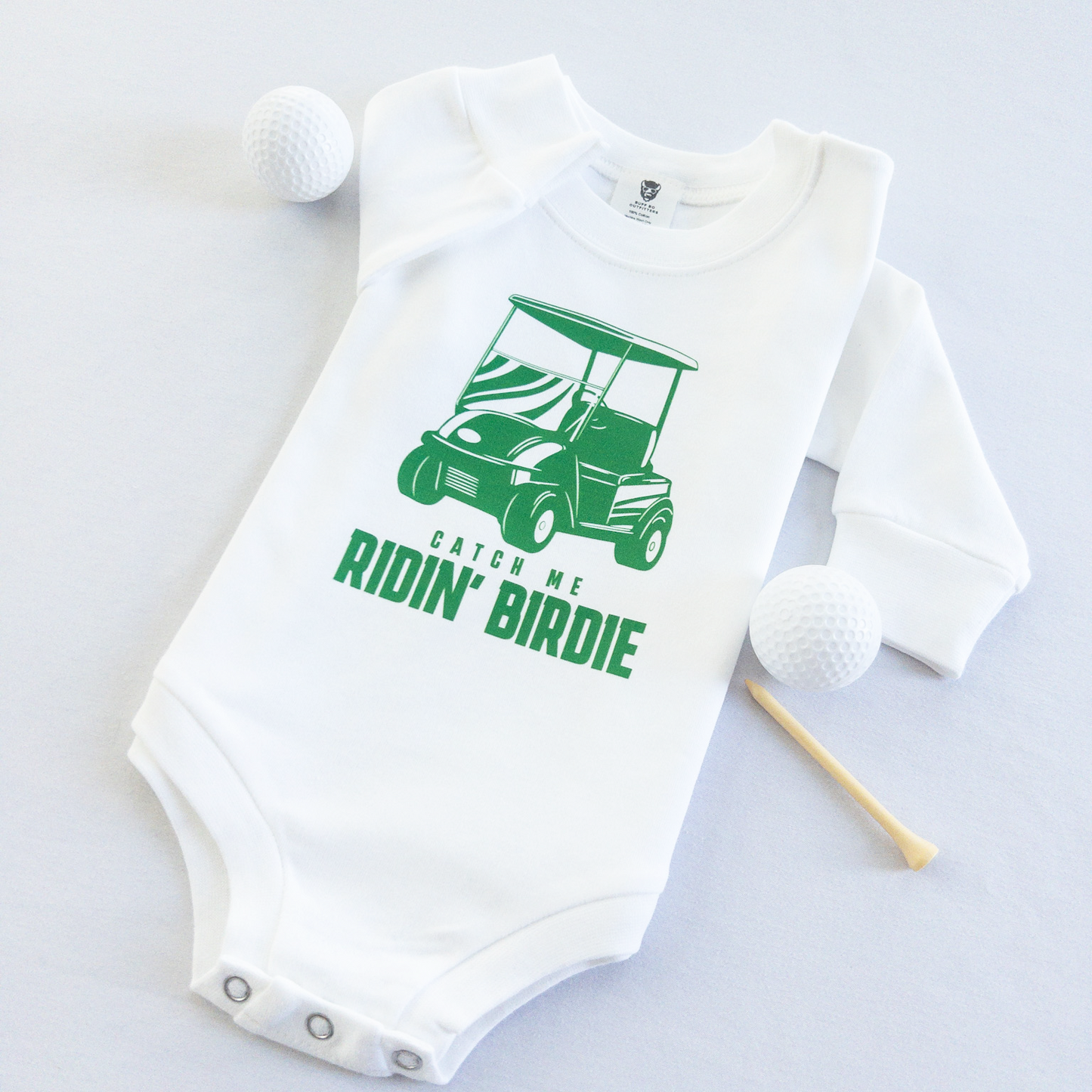 White baby golf onesie featuring a green golf cart graphic and playful text reading 'Catch Me Ridin' Birdie,' styled flat with two golf balls and a wooden tee, fun golf-themed outfit for babies and toddlers.