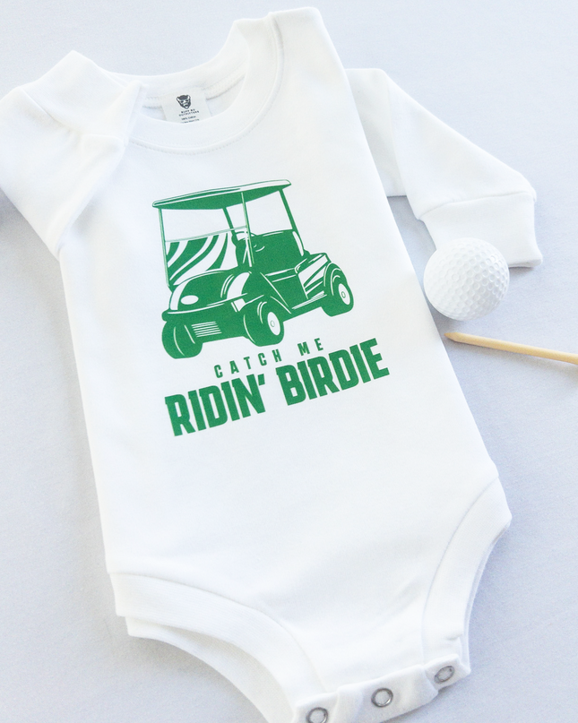 White baby golf onesie featuring a green golf cart graphic and playful text reading 'Catch Me Ridin' Birdie,' styled flat with two golf balls and a wooden tee, fun golf-themed outfit for babies and toddlers.