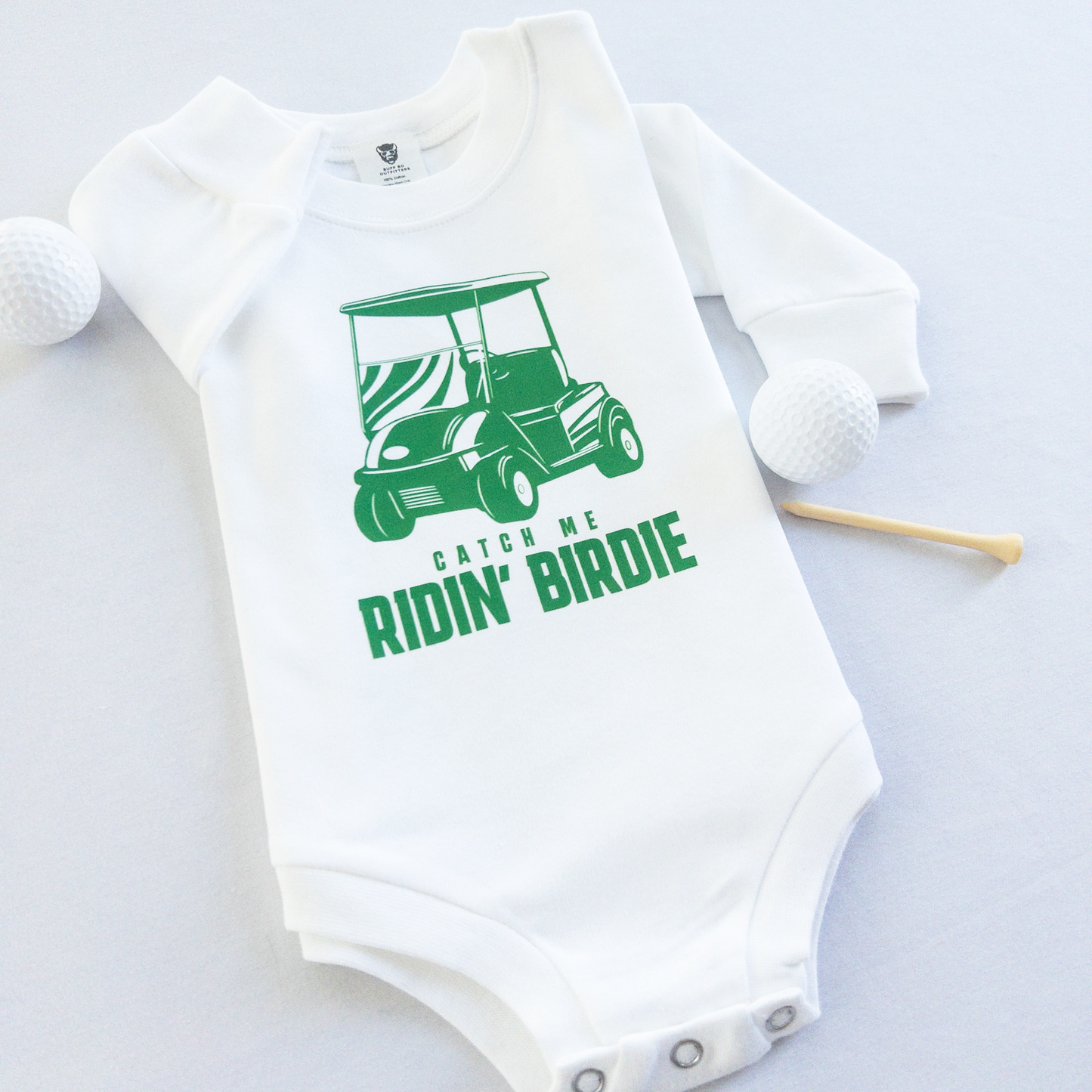 White baby golf onesie featuring a green golf cart graphic and playful text reading 'Catch Me Ridin' Birdie,' styled flat with two golf balls and a wooden tee, fun golf-themed outfit for babies and toddlers.