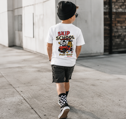 A child wearing a white graphic t-shirt with a 'Skip School' cartoon print, black shorts, a black beanie, and patterned socks, walking away on a concrete sidewalk.