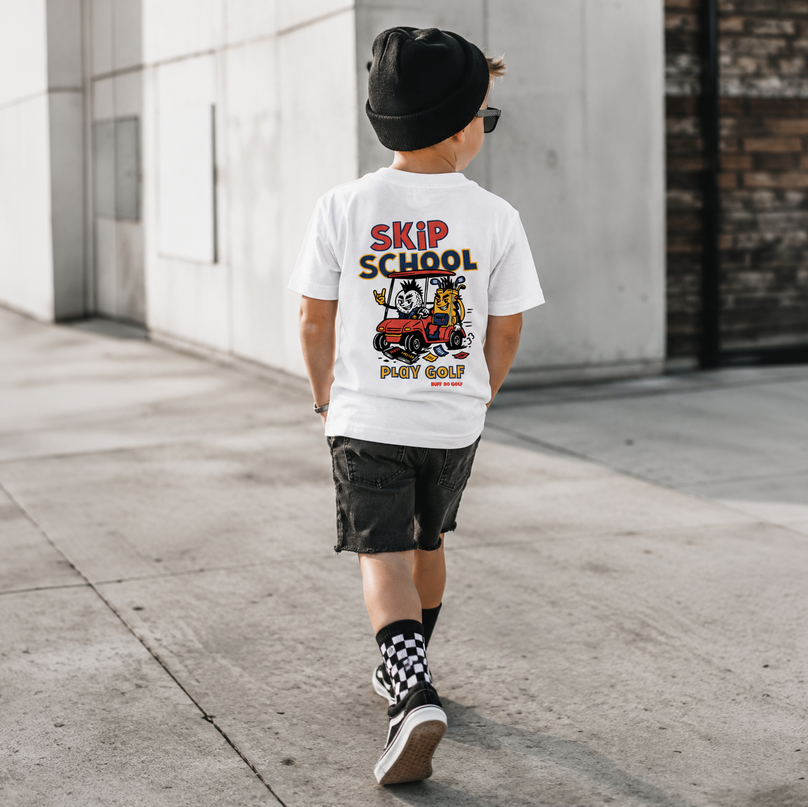 A child wearing a white graphic t-shirt with a 'Skip School' cartoon print, black shorts, a black beanie, and patterned socks, walking away on a concrete sidewalk.