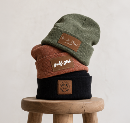 Three knit beanie hats stacked on a wooden stool against a light textured wall. The hats are olive green, rust orange, and black, each with a brown leather patch.