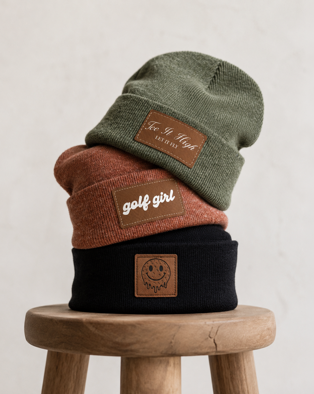 Three knit beanie hats stacked on a wooden stool against a light textured wall. The hats are olive green, rust orange, and black, each with a brown leather patch.