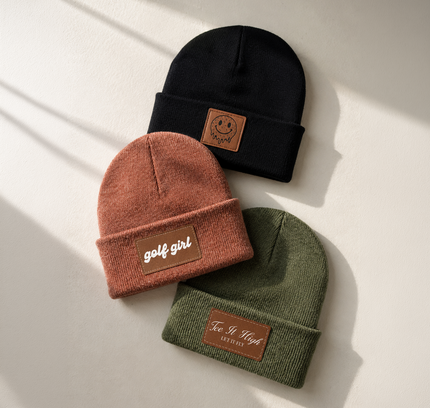 Three knit beanie hats in black, terracotta, and olive green, arranged on a light beige surface with soft diagonal shadows. Each hat has a brown leather patch on the cuff with the words 'golf girl' or 'Fe.