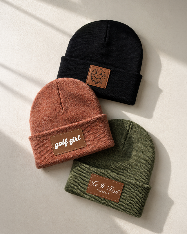 Three knit beanie hats in black, terracotta, and olive green, arranged on a light beige surface with soft diagonal shadows. Each hat has a brown leather patch on the cuff with the words 'golf girl' or 'Fe.