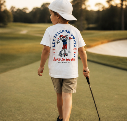 Child on a golf course wearing a white t-shirt with text and graphics, holding a golf club.