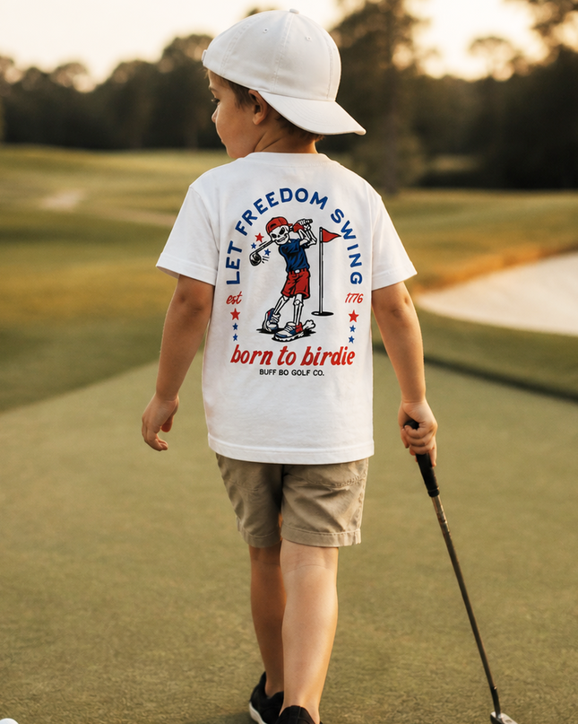 Child on a golf course wearing a white t-shirt with text and graphics, holding a golf club.