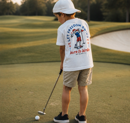 Child on a golf course wearing a white t-shirt with graphics and text.