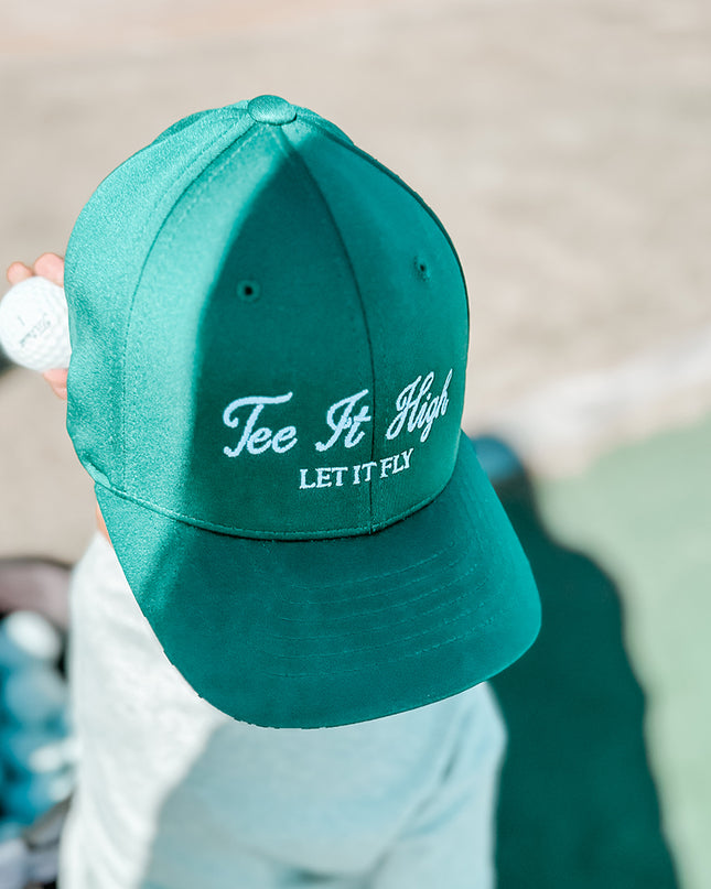 Child holding a golf ball while wearing a green hat with white embroidered text 'Tee It High, Let It Fly,' playful kids golf hat perfect for young golfers, photographed at a golf practice area.