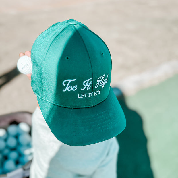 Child holding a golf ball while wearing a green hat with white embroidered text 'Tee It High, Let It Fly,' playful kids golf hat perfect for young golfers, photographed at a golf practice area.