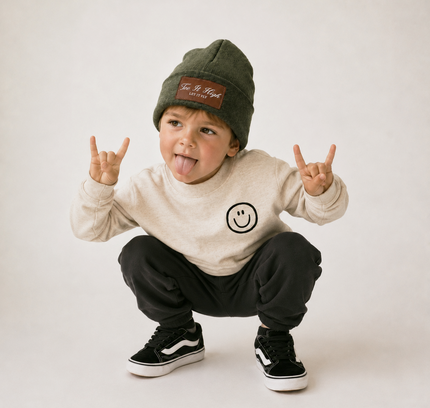 A toddler crouching and sticking out their tongue, wearing an olive green beanie with a brown patch, a cream sweatshirt with a smiley face, black sweatpants, and black low-top sneakers with white stripes, against a plain light background.