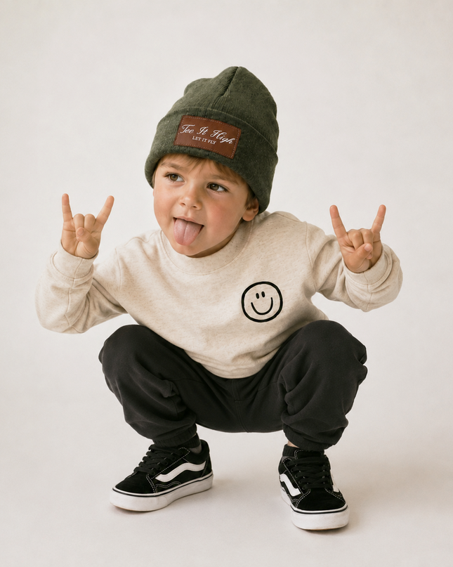 A toddler crouching and sticking out their tongue, wearing an olive green beanie with a brown patch, a cream sweatshirt with a smiley face, black sweatpants, and black low-top sneakers with white stripes, against a plain light background.