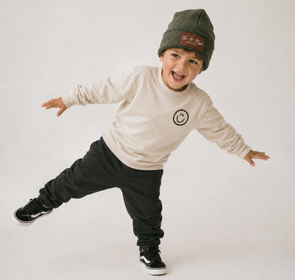 A young boy wearing an olive green beanie with a brown patch, a cream sweatshirt with a smiley face logo, black sweatpants, and black sneakers, posing dynamically against a light gray studio background.