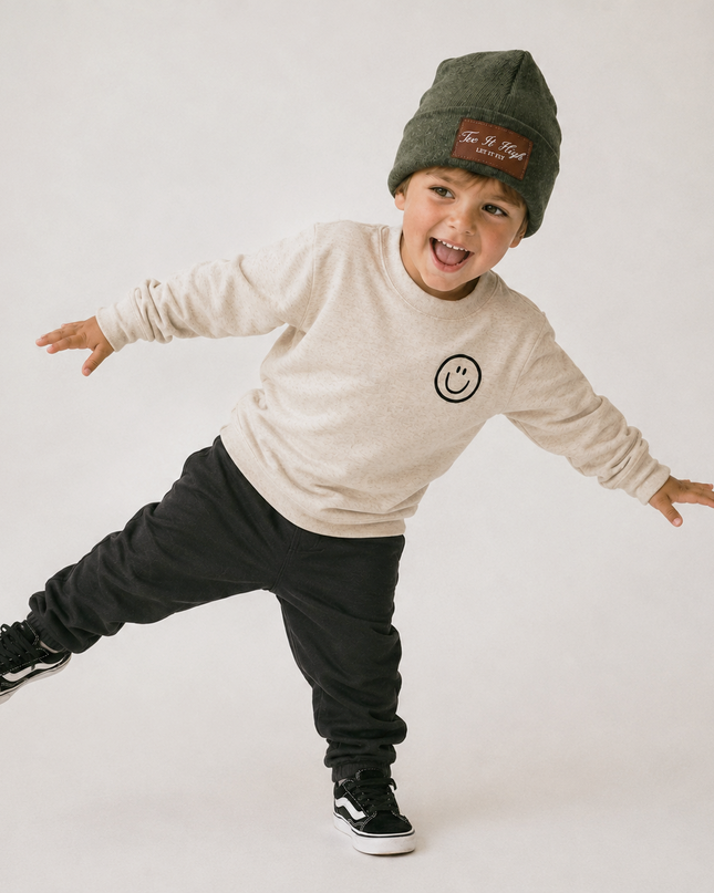 A young boy wearing an olive green beanie with a brown patch, a cream sweatshirt with a smiley face logo, black sweatpants, and black sneakers, posing dynamically against a light gray studio background.