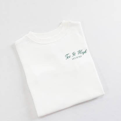 White kids golf t-shirt with minimalist green text on the chest reading 'Tee It High, Let It Fly,' folded neatly on a light background, perfect for young golfers and golf fans.