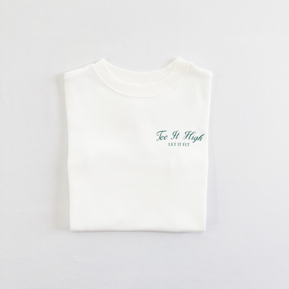 White kids golf t-shirt with minimalist green text on the chest reading 'Tee It High, Let It Fly,' folded neatly on a light background, perfect for young golfers and golf fans.