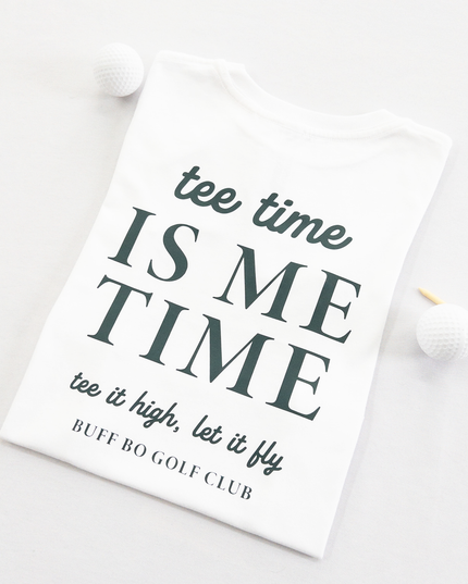 White kids golf t-shirt featuring dark green text reading 'tee time is me time' with additional script text 'tee it high, let it fly' and 'Buff Bo Golf Club,' styled flat with golf balls and a wooden tee, fun and motivational golf apparel for kids.