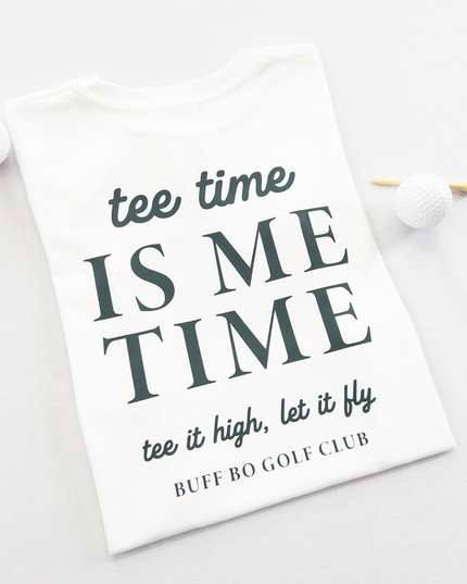 White kids golf t-shirt featuring dark green text reading 'tee time is me time' with additional script text 'tee it high, let it fly' and 'Buff Bo Golf Club,' styled flat with golf balls and a wooden tee, fun and motivational golf apparel for kids.