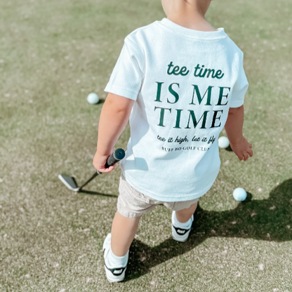 Toddler standing on a putting green wearing a white Buff Bo Outfitters t-shirt that reads “tee time IS ME TIME” and holding a golf club, surrounded by several golf balls.