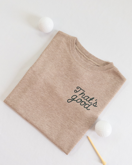Tan kids golf t-shirt featuring dark green script text reading 'That's good,' styled flat lay with two golf balls and a wooden tee, playful and casual golf apparel for young players.