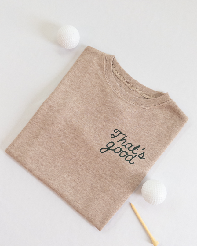 Tan kids golf t-shirt featuring dark green script text reading 'That's good,' styled flat lay with two golf balls and a wooden tee, playful and casual golf apparel for young players.