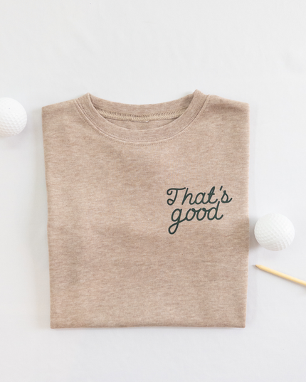 Tan kids golf t-shirt featuring dark green script text reading 'That's good,' styled flat lay with two golf balls and a wooden tee, playful and casual golf apparel for young players.