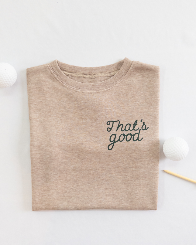 Tan kids golf t-shirt featuring dark green script text reading 'That's good,' styled flat lay with two golf balls and a wooden tee, playful and casual golf apparel for young players.