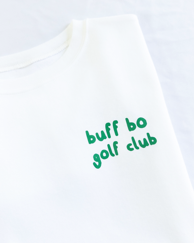 Folded white t-shirt with small green text on the front that reads 'buff bo golf club,' placed next to a white golf ball and wooden tee on a light background.