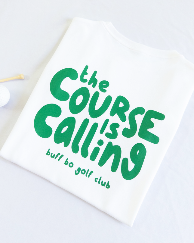 Folded white t-shirt with green text that reads 'the course is calling, buff bo golf club,' next to a white golf ball and wooden tee on a light background.
