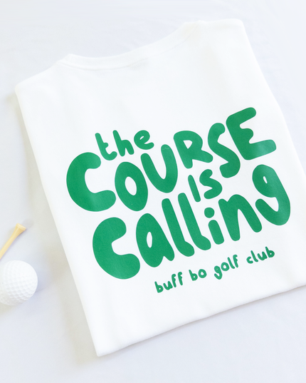 Folded white t-shirt with green text that reads 'the course is calling, buff bo golf club,' next to a white golf ball and wooden tee on a light background.
