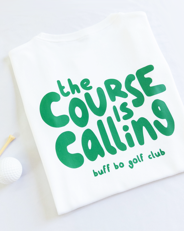 Folded white t-shirt with green text that reads 'the course is calling, buff bo golf club,' next to a white golf ball and wooden tee on a light background.
