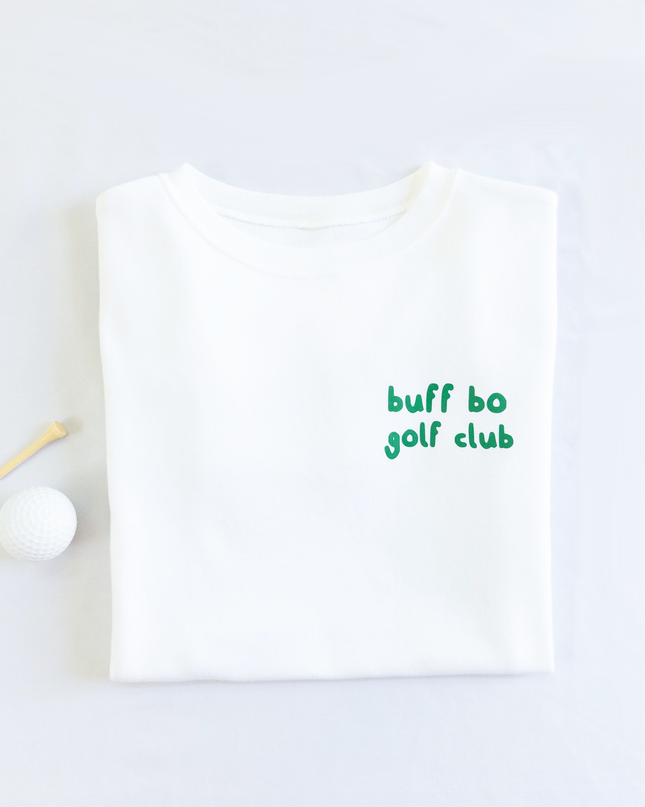 Folded white t-shirt with small green text on the front that reads 'buff bo golf club,' placed next to a white golf ball and wooden tee on a light background.