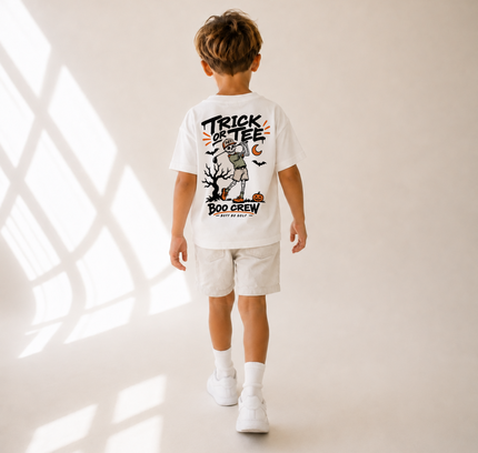 A child model viewed from behind wearing a white graphic t-shirt and light beige shorts in a brightly lit studio with window shadow patterns on the floor.