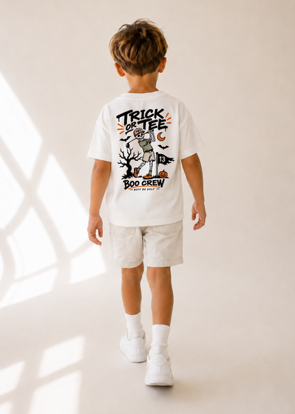 A child viewed from behind wearing a white graphic T-shirt and light shorts, standing in a warm neutral studio setting with window shadow patterns on the floor.