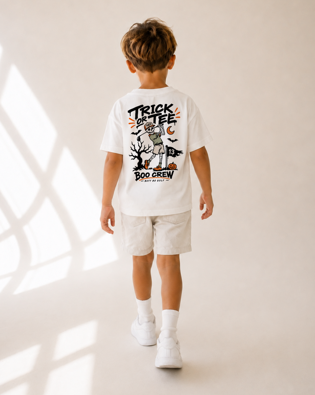 A child viewed from behind wearing a white graphic T-shirt and light shorts, standing in a warm neutral studio setting with window shadow patterns on the floor.