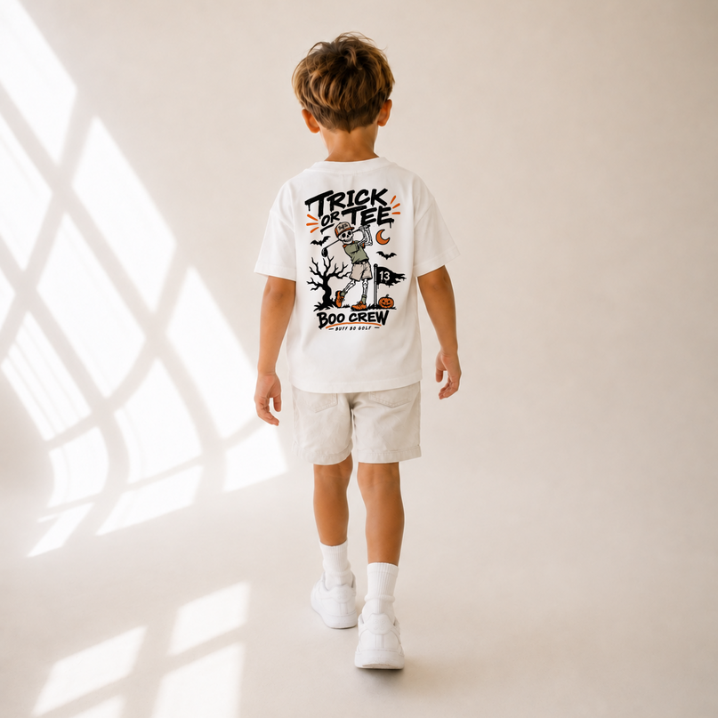 A child viewed from behind wearing a white graphic T-shirt and light shorts, standing in a warm neutral studio setting with window shadow patterns on the floor.