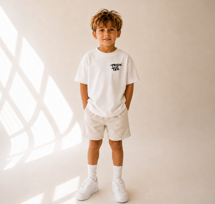 A young boy stands in a studio wearing a white short-sleeve t-shirt with a black 'THINK TREE' logo on the chest, paired with beige shorts, white socks, and white sneakers. The background is a warm beige wall with soft window shadow patterns.
