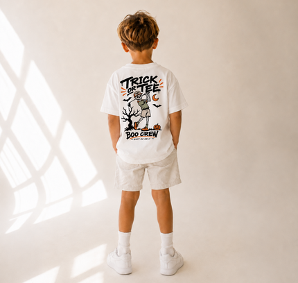 A child standing with their back to the camera, wearing a white graphic t-shirt with a 'Trick or Tee Boo Crew' Halloween design, white shorts, white socks, and white sneakers in a brightly lit studio with window shadow patterns.
