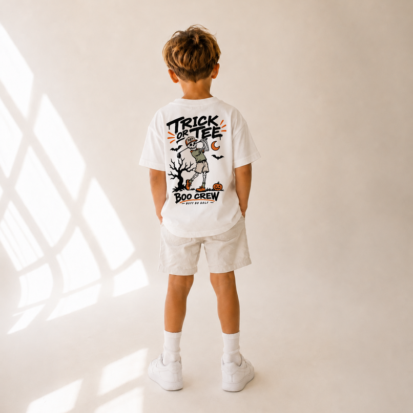 A child standing with their back to the camera, wearing a white graphic t-shirt with a 'Trick or Tee Boo Crew' Halloween design, white shorts, white socks, and white sneakers in a brightly lit studio with window shadow patterns.