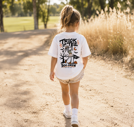 A young girl walking away on a sunlit dirt path, wearing a white graphic T-shirt with a Halloween-themed back print that reads “TRICK OR TEE BOO CREW.”.