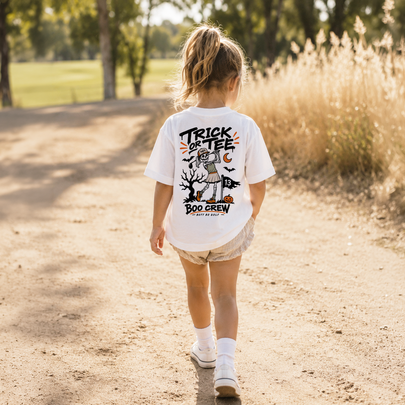 A young girl walking away on a sunlit dirt path, wearing a white graphic T-shirt with a Halloween-themed back print that reads “TRICK OR TEE BOO CREW.”.