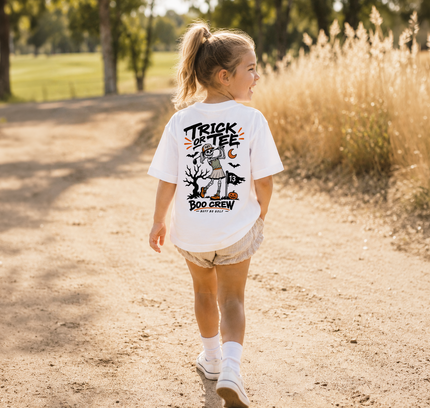 A young girl walking on a sunlit dirt path outdoors, viewed from behind, wearing a white graphic T-shirt with a Halloween-themed design and the words 'TRICK OR TEE' and 'BOO GREW'.
