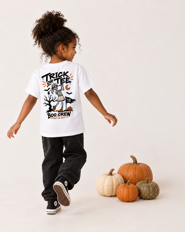 A young child wearing a white graphic t-shirt, black pants, and black sneakers is shown from behind, walking away from the camera. The child is positioned next to a small pile of decorative pumpkins on a clean, light beige studio background.