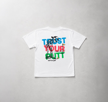 A white short-sleeve t-shirt laid flat on a light gray textured background, featuring a colorful graphic print on the back with the text 'TRUST YOUR BUTT' in blue, red, and green, and a cartoon character wearing sunglasses.