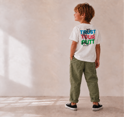 A child stands with their back to the camera wearing a white t-shirt with a colorful graphic that reads 'TRUST YOUR GUT' and olive green pants, set against a warm beige textured wall with soft window light shadows.