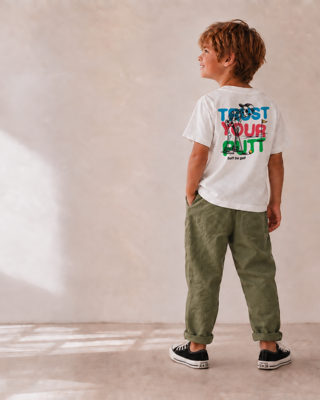 A child stands with their back to the camera wearing a white t-shirt with a colorful graphic that reads 'TRUST YOUR GUT' and olive green pants, set against a warm beige textured wall with soft window light shadows.